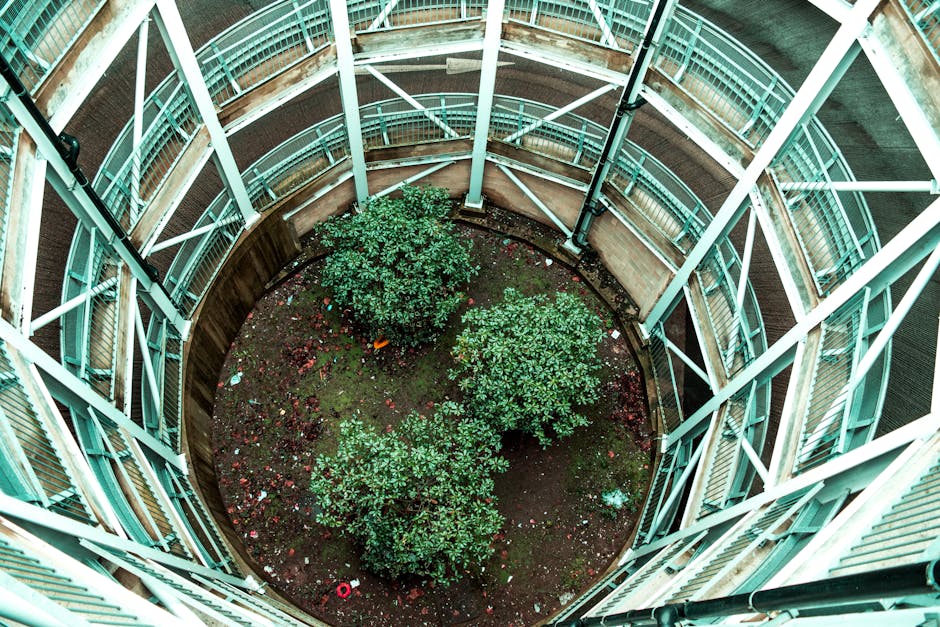 An overhead view of a circular garden courtyard enclosed by a multi-story building with turquoise metal railings and glass balconies. The garden features three lush green bushes evenly spaced within a