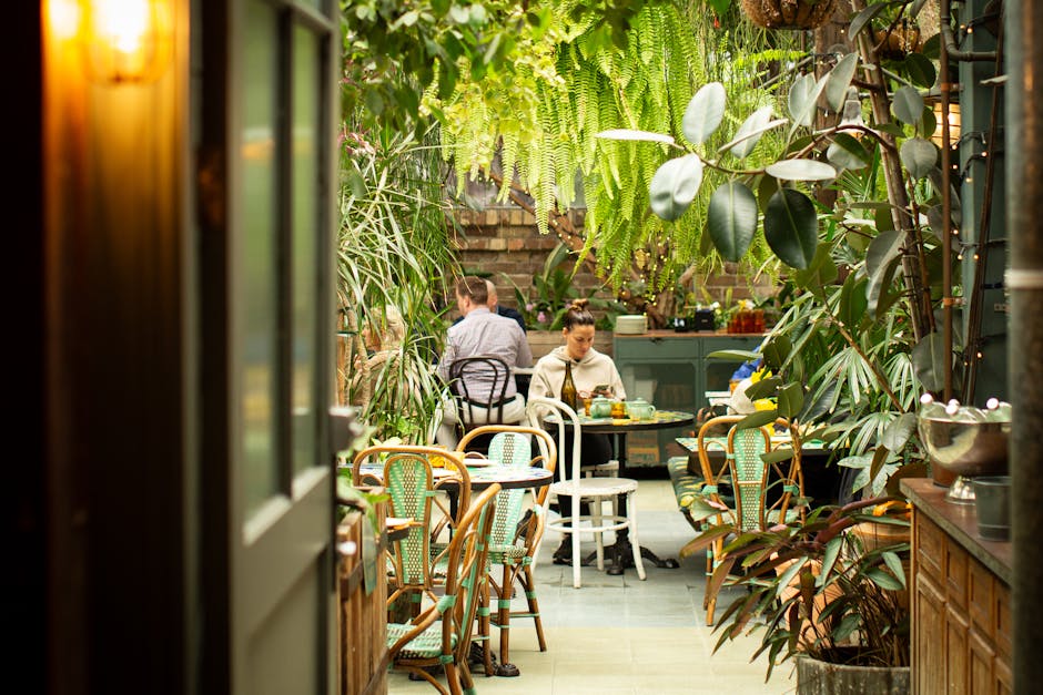 An overhead view of a circular garden courtyard enclosed by a multi-story building with turquoise metal railings and glass balconies. The garden features three lush green bushes evenly spaced within a